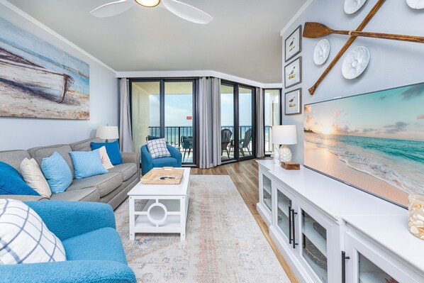A living room with a sofa, coffee table, and a TV displaying a beach scene. - Phoenix Iii 3035 1 Bedroom Condo (Orange Beach)