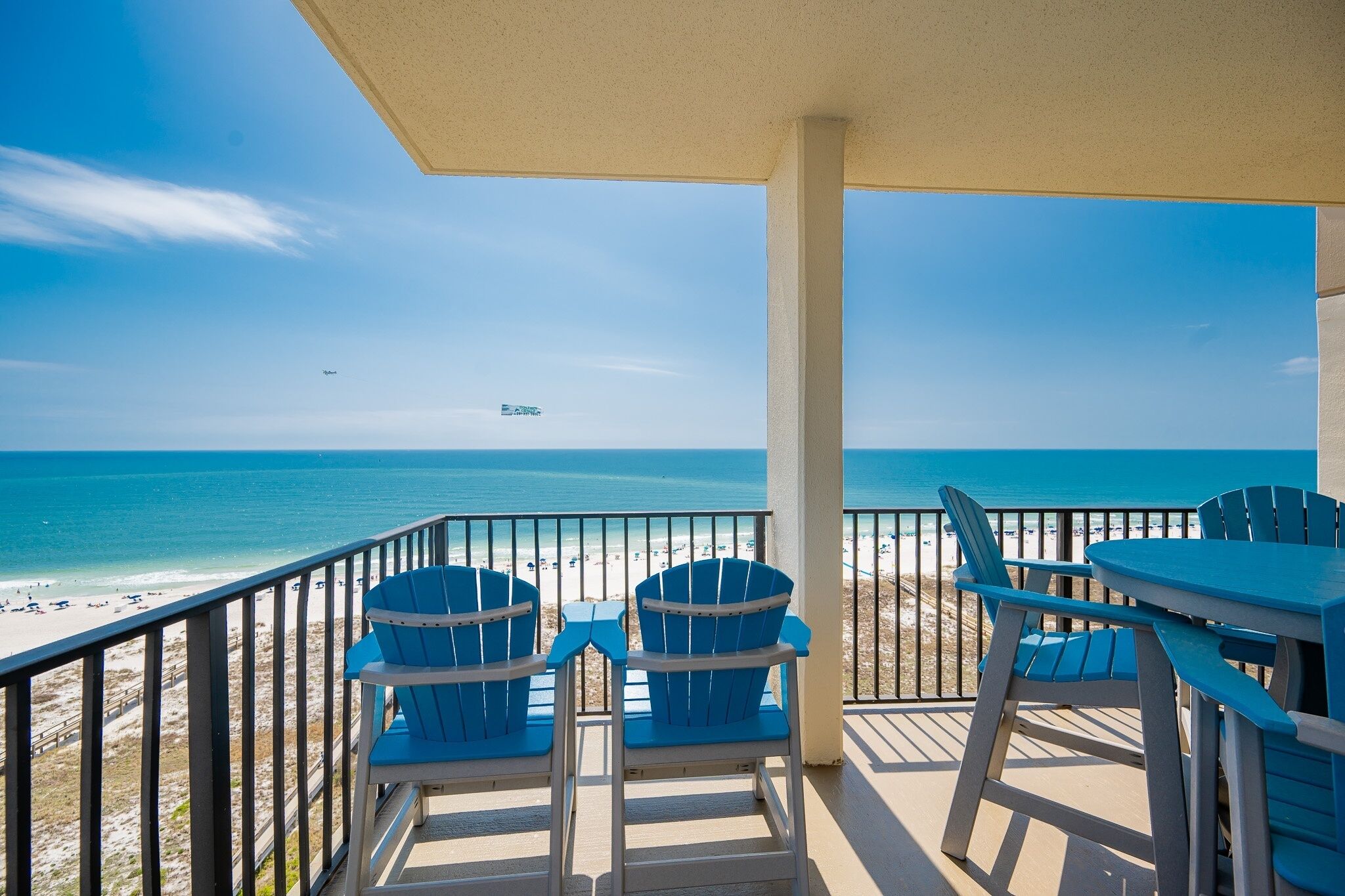 A balcony with a view of the beach and ocean, featuring two blue chairs and a table.