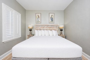 A bedroom with a large bed, two bedside tables with lamps, a window with blinds, and two wall-mounted paintings. - Phoenix Vii 71517 3 Bedroom Condo (Orange Beach)