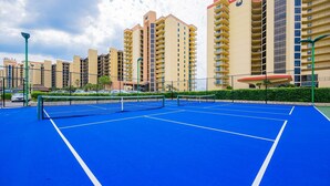 A tennis court with a net, surrounded by a fence, and adjacent to residential buildings. - Phoenix Iii 3057 1 Bedroom Condo (Orange Beach)