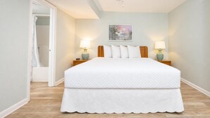 A bedroom with a bed, two bedside tables with lamps, a door leading to a bathroom, and a painting on the wall. - Phoenix Iii 3057 1 Bedroom Condo (Orange Beach)