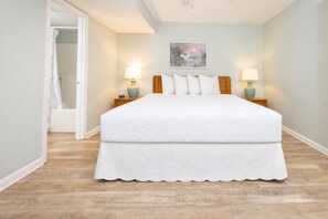 A bedroom with a bed, two bedside tables with lamps, a door leading to a bathroom, and a painting on the wall. - Phoenix Iii 3057 1 Bedroom Condo (Orange Beach)