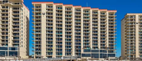 Un hotel moderno de gran altura con varios balcones, una playa arenosa al frente y un cielo azul despejado.