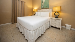 A bedroom with a large bed, two bedside tables with lamps, a painting on the wall, and a curtain on the window. - Phoenix Vii 7103 3 Bedroom Condo (Orange Beach)