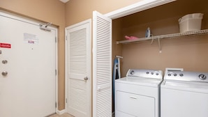 A laundry room with two stacked washers and dryers, a metal shelf with laundry supplies, and a door with a safety sign. - Phoenix Vii 7103 3 Bedroom Condo (Orange Beach)