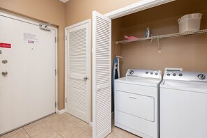 A laundry room with two stacked washers and dryers, a metal shelf with laundry supplies, and a door with a safety sign. - Phoenix Vii 7103 3 Bedroom Condo (Orange Beach)