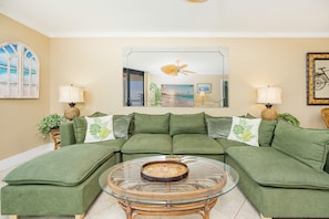A green sectional sofa with a glass coffee table in the center, a ceiling fan, and a large mirror above the sofa. - Phoenix Vii 71010 2 Bedroom Condo (Orange Beach)