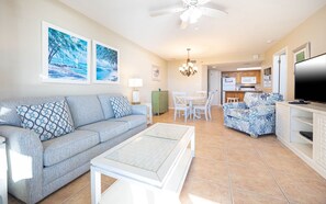 Ein Wohnzimmer mit einer Couch, einem Couchtisch und einem Fernseher. Es gibt einen Essbereich mit Stühlen und einem Tisch. Der Raum verfügt über einen Deckenventilator und Kunstwerke an den Wänden.