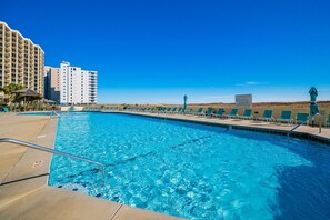 A swimming pool with clear blue water, surrounded by lounge chairs and a building with multiple floors. - Phoenix Vii 71506 2 Bedroom Condo (Orange Beach)