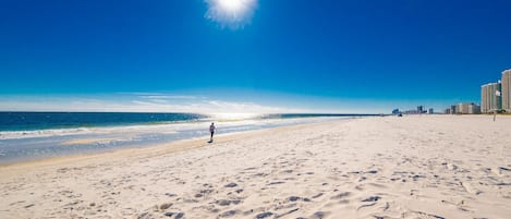 Una amplia playa de arena con huellas, cielo azul despejado y un horizonte con edificios de la ciudad a lo lejos.