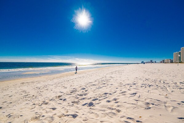 A wide sandy beach with footprints, a clear blue sky, and a distant cityscape. - Phoenix Vii 71506 2 Bedroom Condo (Orange Beach)