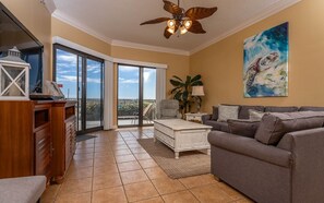 Ein Wohnzimmer mit grauem Sofa, weißem Couchtisch und Deckenventilator. Ein großes Fenster bietet Blick auf den Himmel, und eine Tür führt zu einem Balkon.