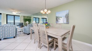 Ruokailutilassa on puinen pöytä ja tuolit, kattotuuletin ja suurista ikkunoista avautuu näkymä merelle.
