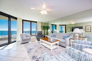 A spacious living room with a view of the beach, a ceiling fan, and a large window. - Phoenix Vii 7608 3 Bedroom Condo (Orange Beach)