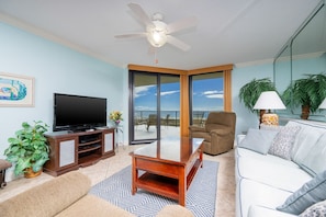 A living room with a flat-screen TV, a wooden entertainment center, a dining table, a sofa, and a balcony with a view. - Phoenix Vii 7309 2 Bedroom Condo (Orange Beach)
