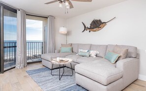 A modern living room with a grey sectional sofa, a small round coffee table, and a view of the ocean through large sliding glass doors. - Phoenix Vii 7813 2 Bedroom Condo (Orange Beach)