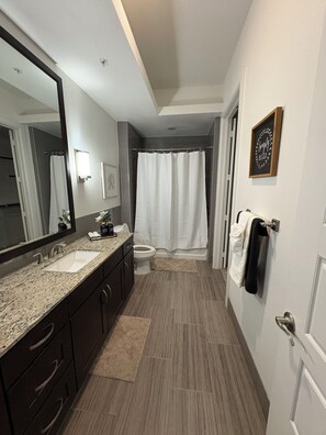 A modern bathroom with a double sink vanity, a large mirror, a shower with a curtain, and a towel rack. - Luxury 2BR • King Suite • Minutes to PGA, Universal & The Star (Frisco)