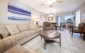 Ein Wohnzimmer mit einer Couch, Sesseln, einem Couchtisch, einem Deckenventilator und einer Küche im Hintergrund.