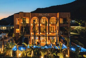 A modern hotel with a pool, palm trees, and a mountainous backdrop.