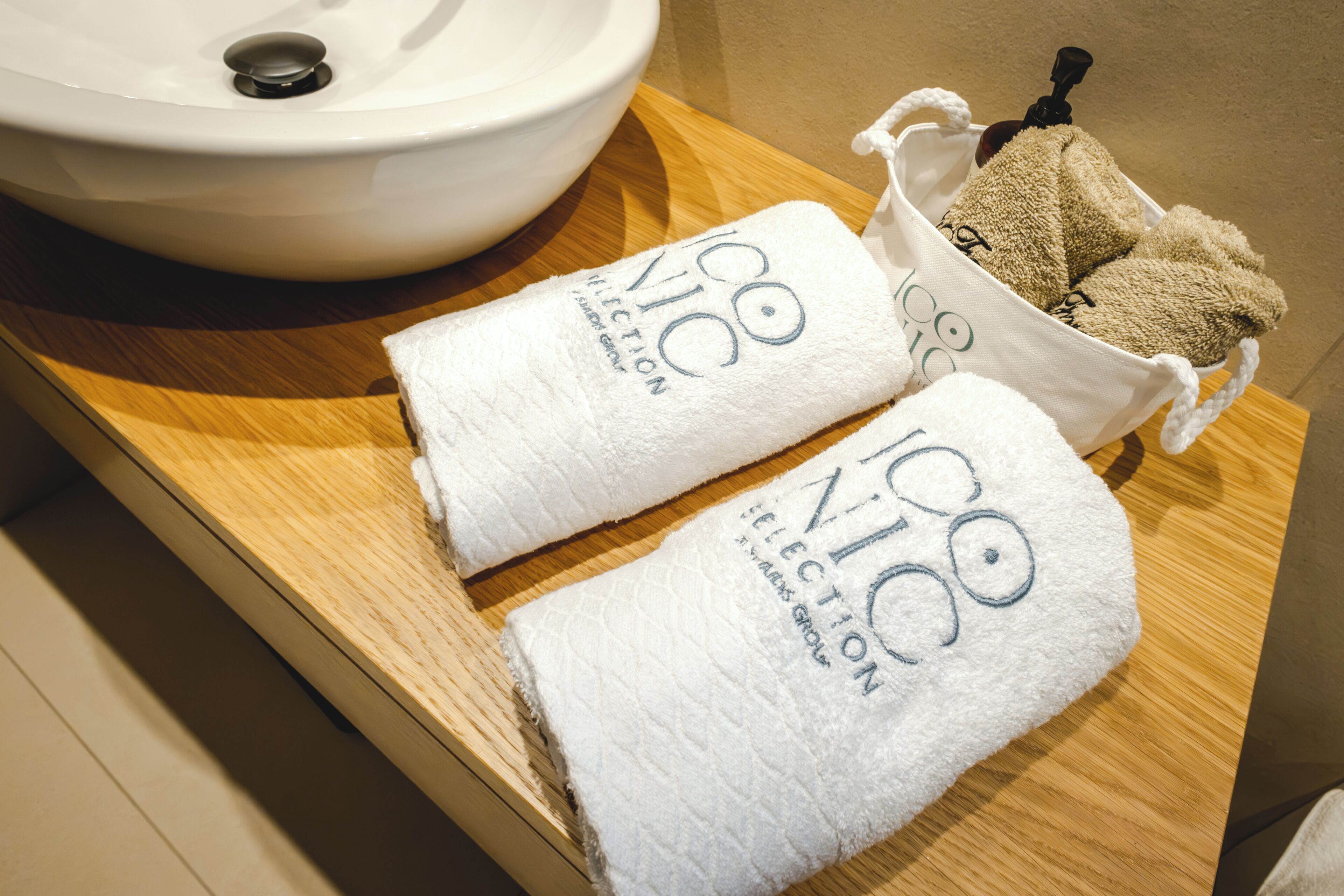 Un baño con un lavabo blanco, dos toallas blancas y una bolsa para toallas color beige sobre una encimera de madera.