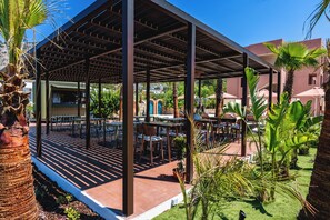 Un área de estar al aire libre cubierta con mesas y sillas, rodeada de follaje y un edificio al fondo.