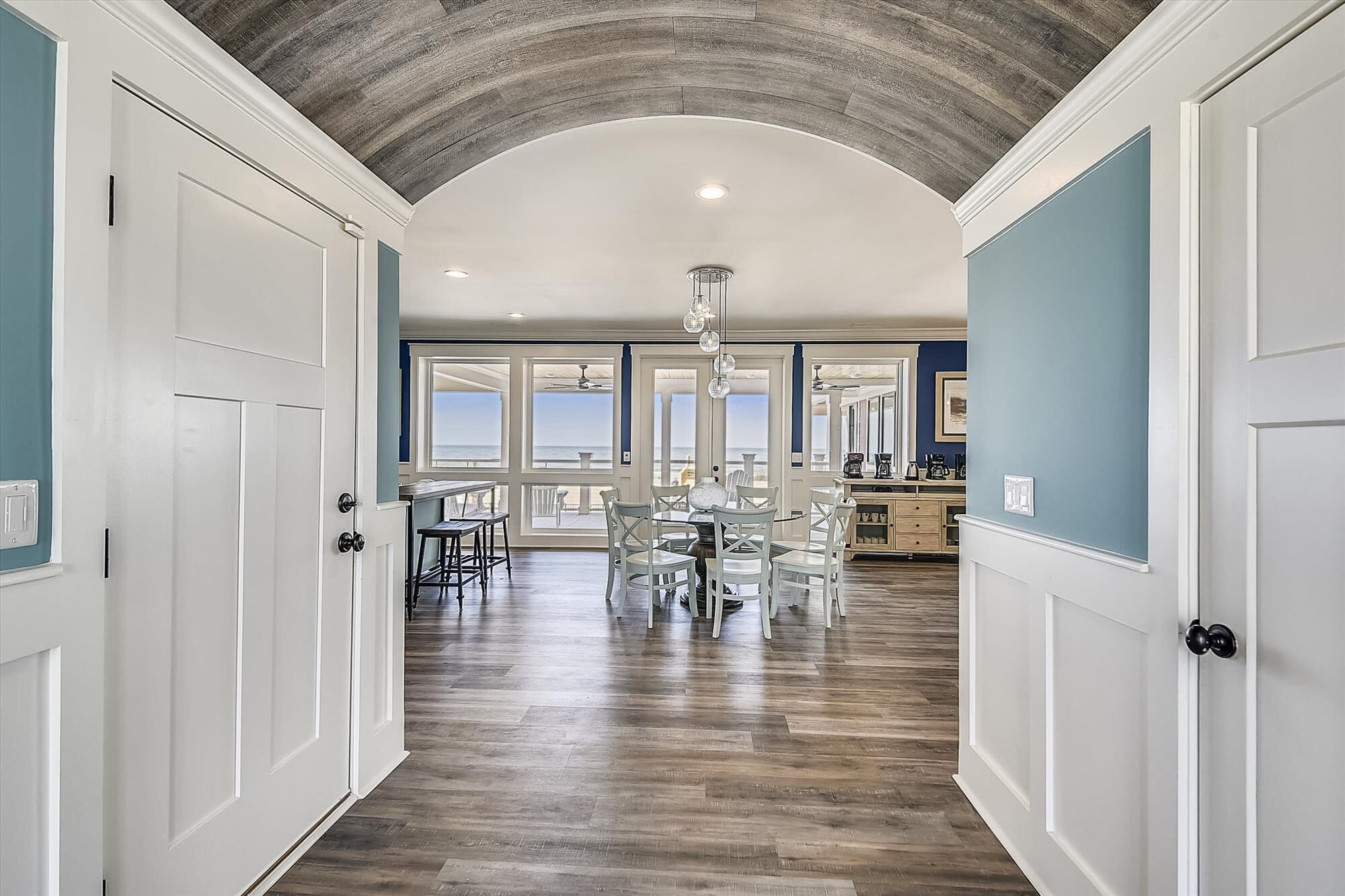A hallway with a wooden floor, white doors, and a view of the ocean through large windows.