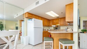 A modern kitchen with wooden cabinets, a white refrigerator, a built-in island with stools, and a tiled floor. - Phoenix Vii 7211 1 Bedroom Condo (Orange Beach)