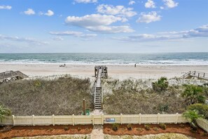 A beach view with a wooden boardwalk leading to the ocean, a white picket fence, and a warning sign. - Majestic Dream: Oceanfront, Pool, Hot Tub & Endless Coastal Days (Murrells Inlet)