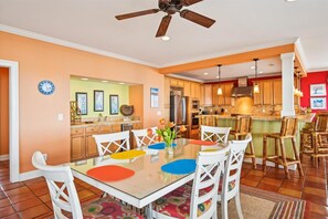 A modern kitchen with wooden cabinets, a stainless steel refrigerator, and a dining area with a glass table and white chairs. - Majestic Dream: Oceanfront, Pool, Hot Tub & Endless Coastal Days (Murrells Inlet)