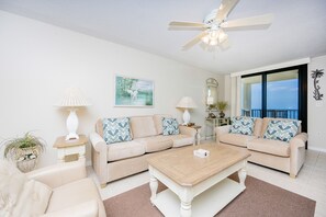 A living room with two beige sofas, a wooden coffee table, a ceiling fan, and a large window with a view. - Phoenix Vii 7602 1 Bedroom Condo (Orange Beach)