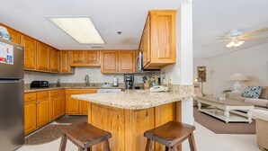 A kitchen with wooden cabinets, a refrigerator, a coffee maker, and a bar stool. - Phoenix Vii 7602 1 Bedroom Condo (Orange Beach)