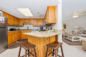 A kitchen with wooden cabinets, a refrigerator, a coffee maker, and a bar stool. - Phoenix Vii 7602 1 Bedroom Condo (Orange Beach)