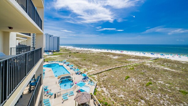 A pool area with multiple swimming pools and lounge chairs, overlooking a beach with a sandy shore and a clear blue sky. - Phoenix Vii 7602 1 Bedroom Condo (Orange Beach)
