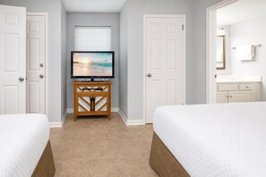 A hotel room with two beds, a television on a wooden stand, a window with blinds, and a bathroom with a mirror and towel rack. - Phoenix Vii 7804 2 Bedroom Condo (Orange Beach)