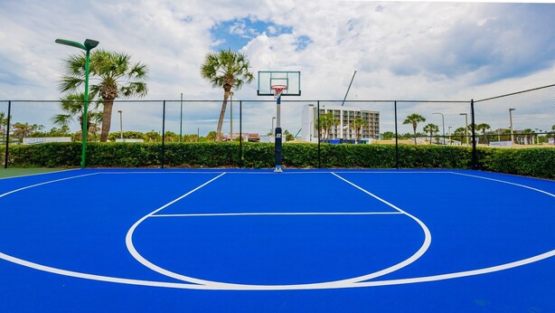 An outdoor basketball court with a blue surface, surrounded by a black fence, palm trees, and a building in the background. - Phoenix Iii 3022 1 Bedroom Condo (Orange Beach)