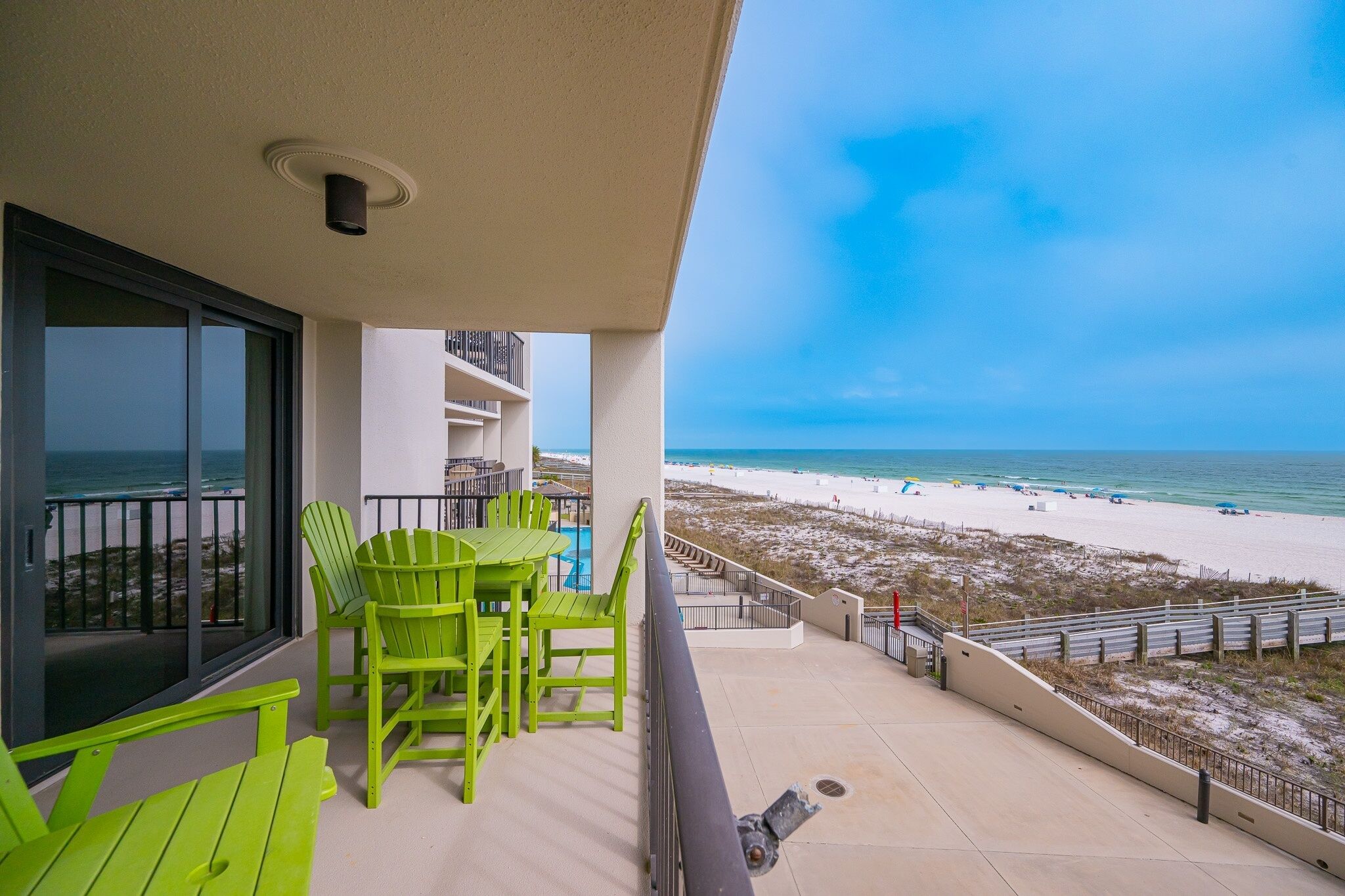 A balcony with green chairs and a table, overlooking a beach with a clear sky.