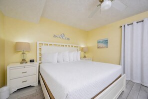 A hotel room with a bed, two bedside tables with lamps, a ceiling fan, a painting on the wall, and a window with curtains. - Phoenix Iii 3022 1 Bedroom Condo (Orange Beach)