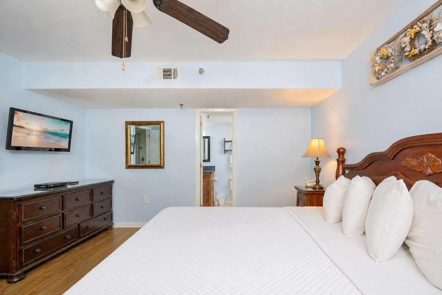 A hotel room with a bed, a TV, a dresser, a ceiling fan, a mirror, and a nightstand with a lamp.
