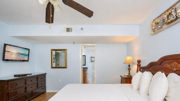 Habitación de hotel con una cama, televisión, cómoda, ventilador de techo, espejo y mesita de noche con lámpara.