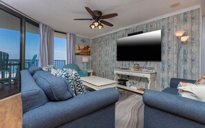 A room with a large TV, a ceiling fan, a dining table, and a view of the ocean. - Phoenix Vii 7917 3 Bedroom Condo (Orange Beach)