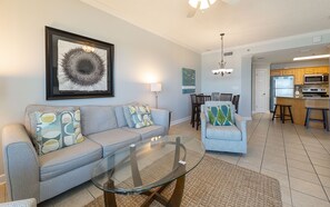 A living room with a sofa, armchair, glass coffee table, dining area with chairs, and a kitchen with appliances. - Phoenix Vii 71215 2 Bedroom Condo (Orange Beach)