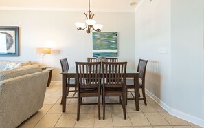 A dining area with a table and chairs, a chandelier, a painting, and a sofa. - Phoenix Vii 71215 2 Bedroom Condo (Orange Beach)