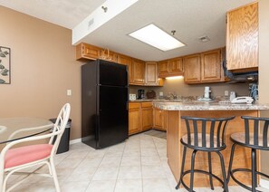 A compact kitchen with wooden cabinets, a black refrigerator, a granite countertop, and a small dining area with a white chair and a red cushion. - Phoenix Iii 3075 1 Bedroom Condo (Orange Beach)