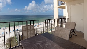 A balcony with a table and chairs overlooking a beach with umbrellas and a clear view of the ocean. - Phoenix Iii 3075 1 Bedroom Condo (Orange Beach)