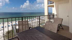 A balcony with a table and chairs overlooking a beach with umbrellas and a clear view of the ocean. - Phoenix Iii 3075 1 Bedroom Condo (Orange Beach)