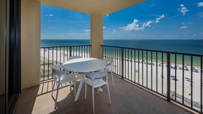Ein Balkon mit Tisch und Stühlen, der Blick auf einen Strand mit Sonnenschirmen und einem klaren Himmel bietet.
