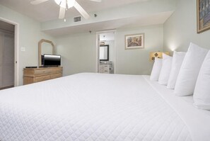 Hotelkamer met groot bed, televisie op een houten dressoir, plafondventilator en een ingelijste foto aan de muur.