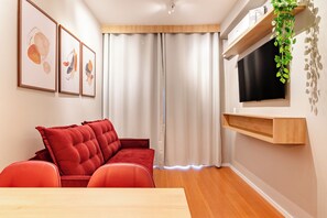 A modern living room with a red sofa, a wooden TV stand, and a large window with curtains. - Maison (São Paulo)