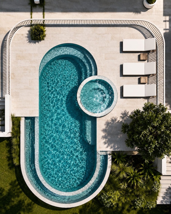 A large oval-shaped pool with a circular hot tub, surrounded by a white deck with lounge chairs, and a small garden area with plants.
