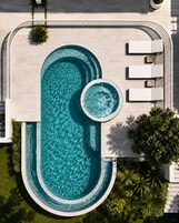 Una piscina ovalada grande con un jacuzzi circular, rodeada por una terraza blanca con sillas de descanso, y un pequeño jardín con plantas.
